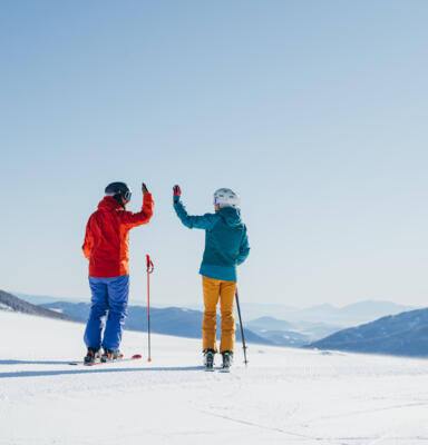 Skifahren_SanktOswald_MBN_ёSamStrauss_Winter2021_28 Zwei Skifahrende stehen auf einer verschneiten Piste und begrüßen sich mit erhobenen Händen.